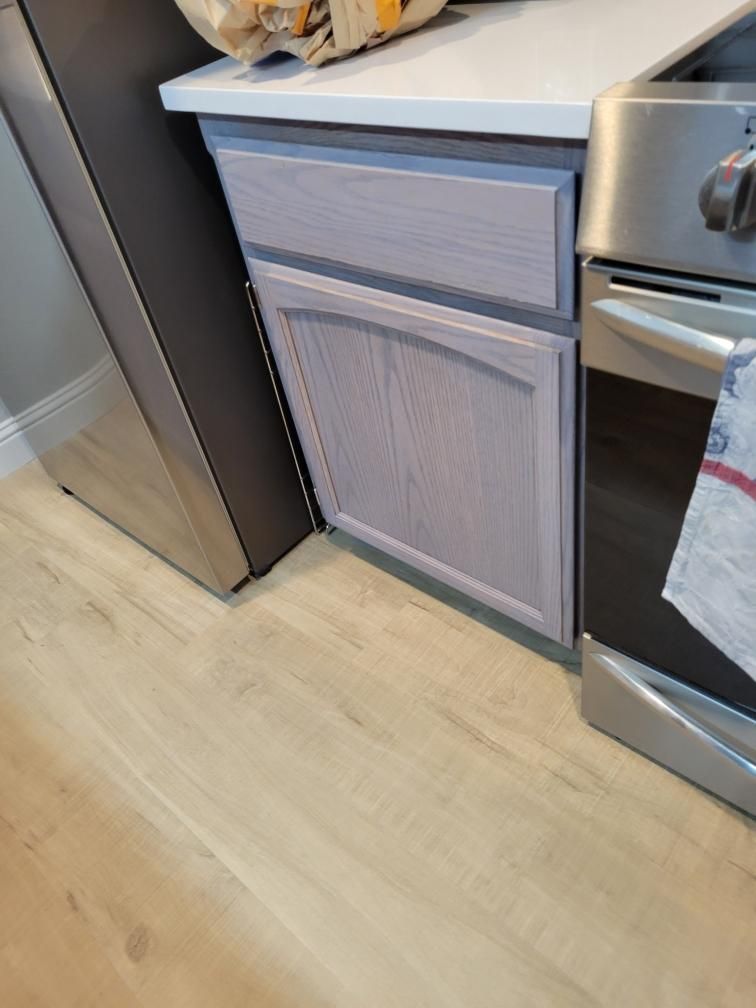 Kitchen cabinet between a stainless refrigerator and oven, with a light wood floor.