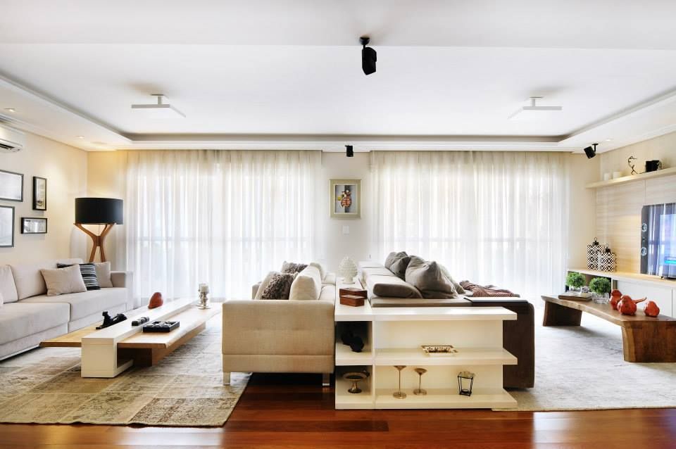 Sala de estar moderna e iluminada com sofás bege, mesas de centro brancas e cortinas translúcidas sobre piso de madeira.