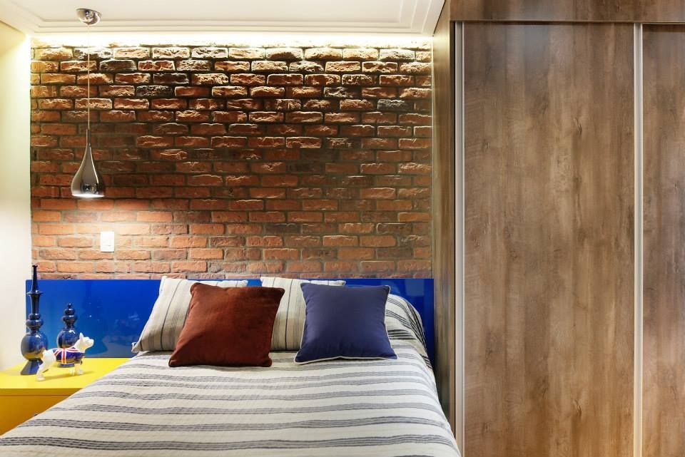 Quarto com parede de tijolos aparentes, guarda-roupa de madeira, cabeceira azul e roupa de cama listrada com travesseiros.