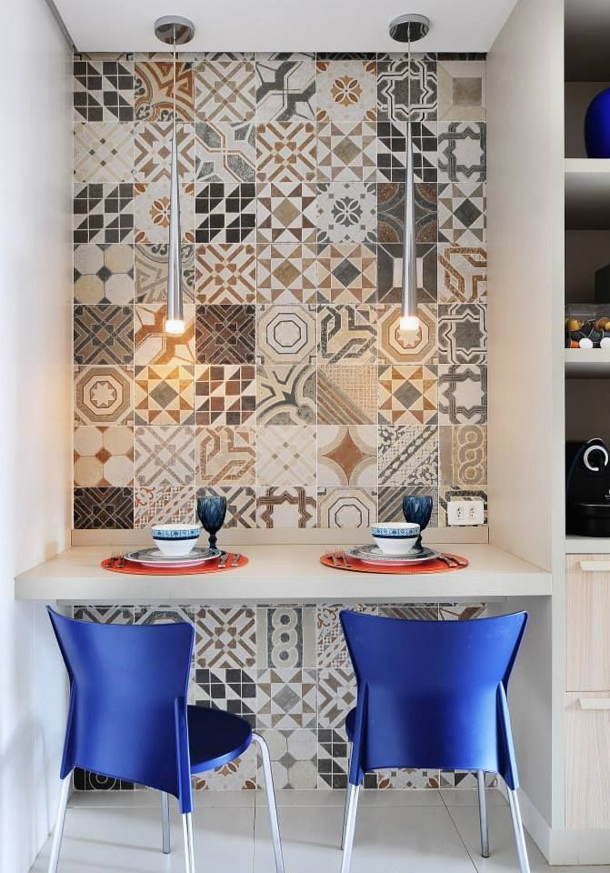 Cantinho de jantar compacto com parede de azulejos estampados, mesa branca e duas cadeiras azuis sob luminárias pendentes.