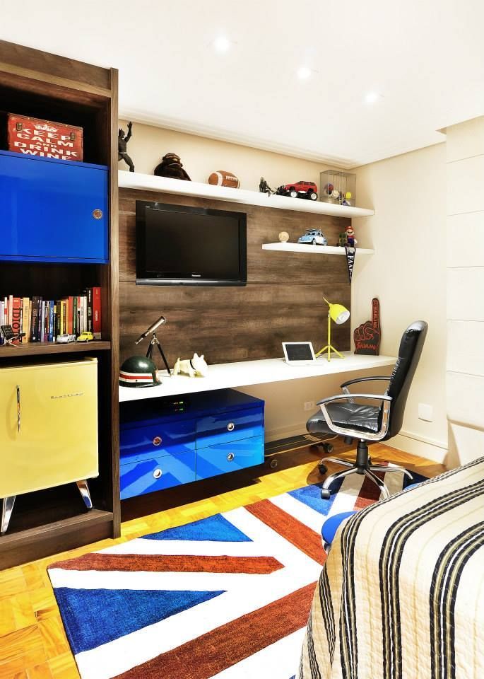 Escritório moderno em casa com mesa, monitor de computador, cadeira preta, prateleiras e tapete geométrico colorido.