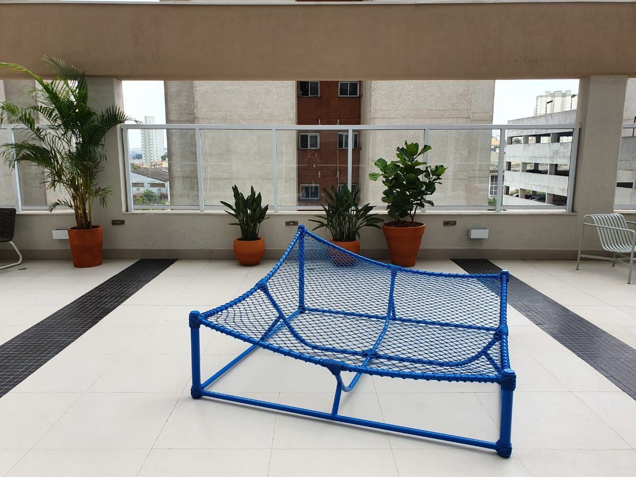 Rede de segurança azul para esportes em um terraço na cobertura, com vasos de plantas e prédios da cidade ao fundo.