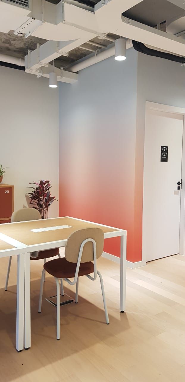 Área de jantar moderna em escritório com mesa de madeira, cadeiras, planta em vaso e porta branca ao lado de uma parede com degradê rosa.