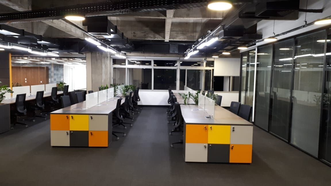 Interior de escritório moderno com fileiras de mesas, cadeiras pretas e armários de armazenamento amarelo-alaranjados sob tubulações expostas no teto.