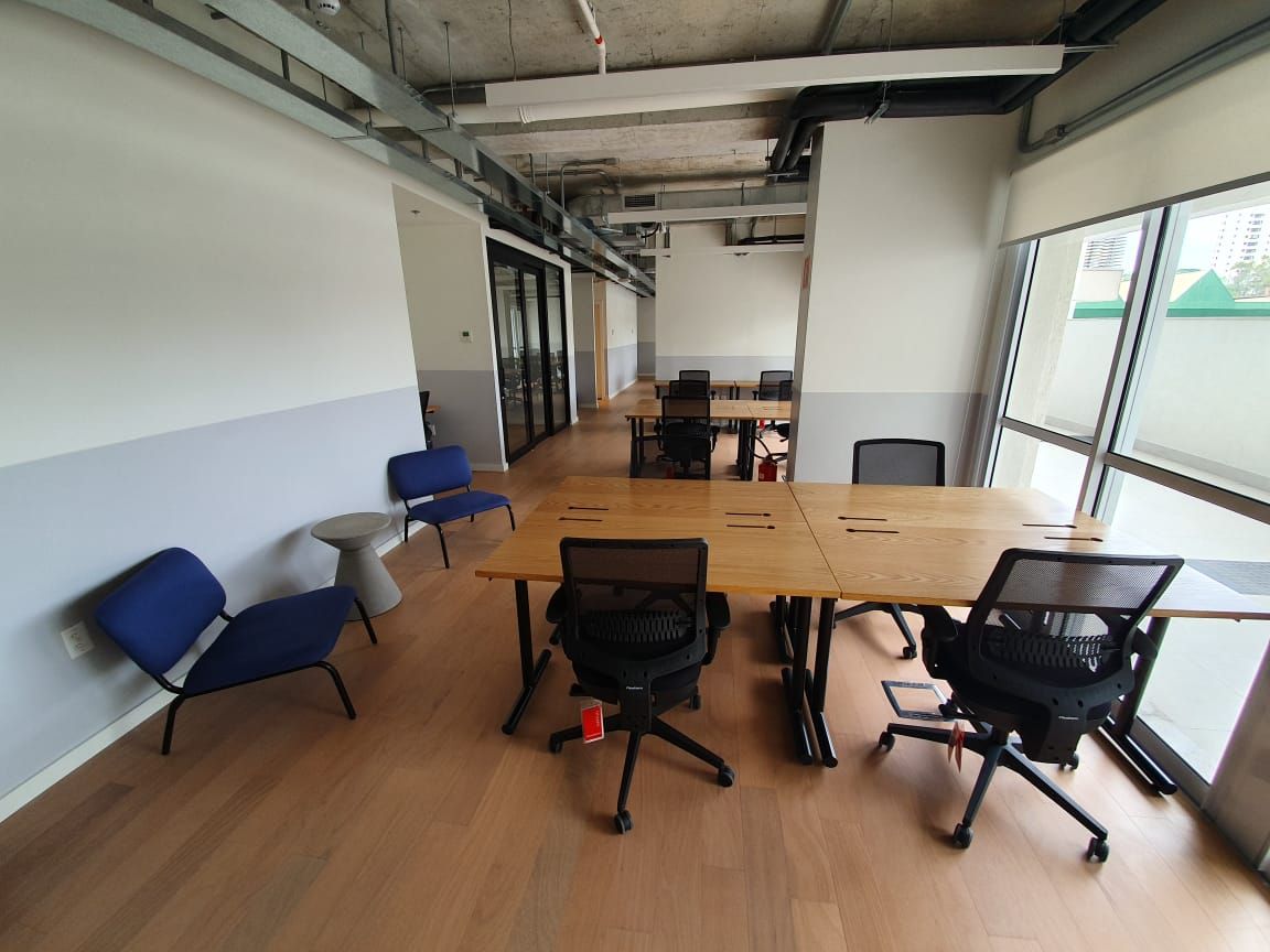 Sala de reuniões vazia com mesa de madeira, cadeiras pretas, poltronas azuis e janelas amplas.