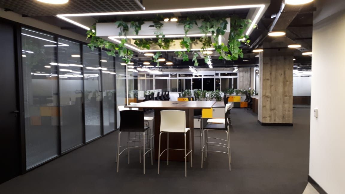 Sala de estar moderna com mesa redonda, cadeiras, paredes de vidro e plantas suspensas sob iluminação intensa.
