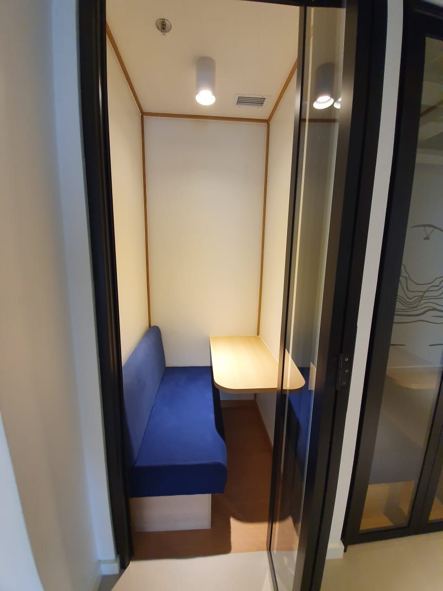 Cabine estreita com banco azul, mesa pequena e luz suspensa em uma sala compacta com paredes de vidro.