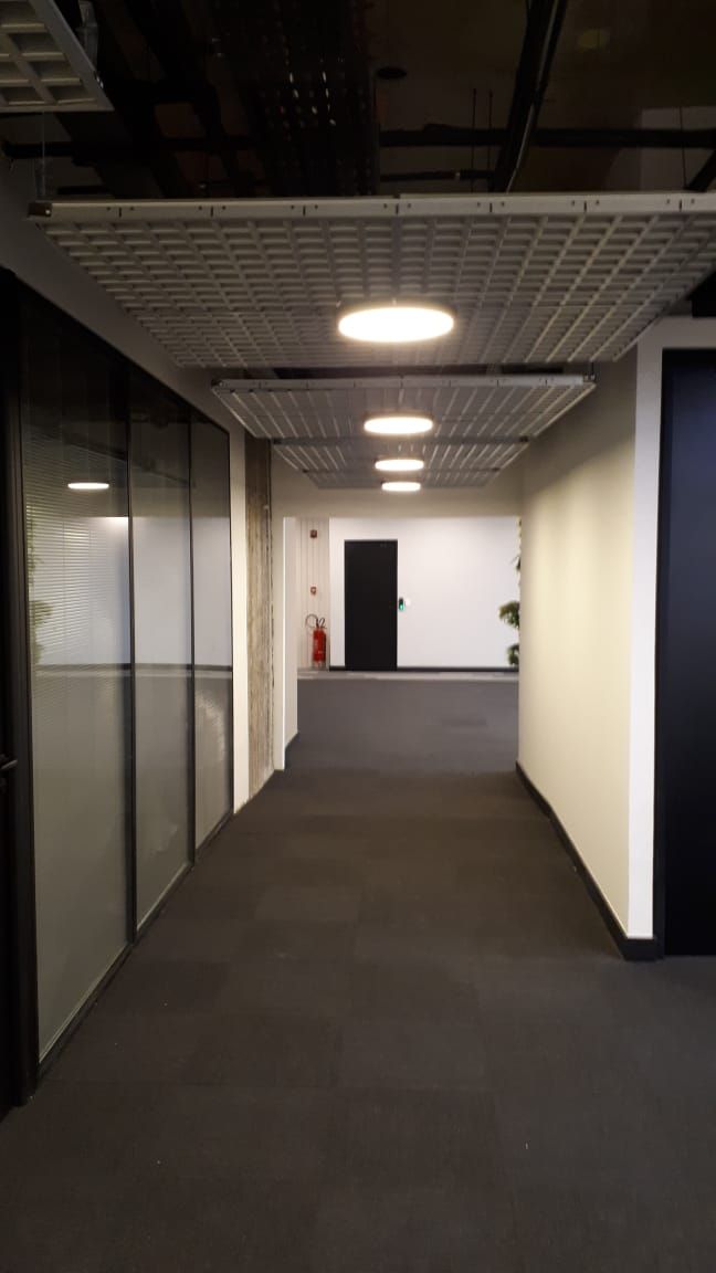 Corredor de escritório vazio com piso acarpetado, paredes de vidro e luzes no teto, que leva a uma porta.
