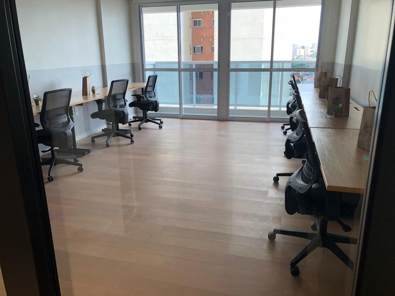 Sala de escritório vazia com mesas, cadeiras com rodinhas e grandes janelas com vista para a cidade.