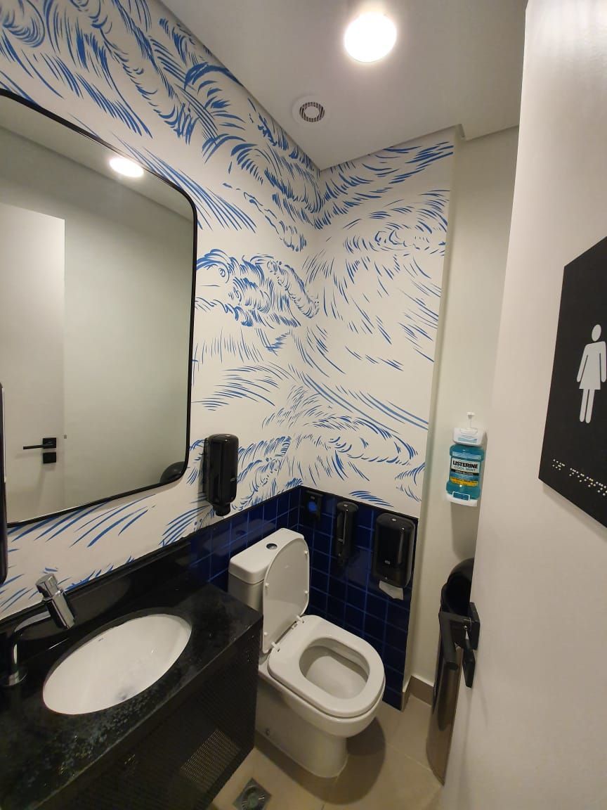 Banheiro pequeno com vaso sanitário, pia, bancada preta, espelho e paredes com estampa azul e branca.