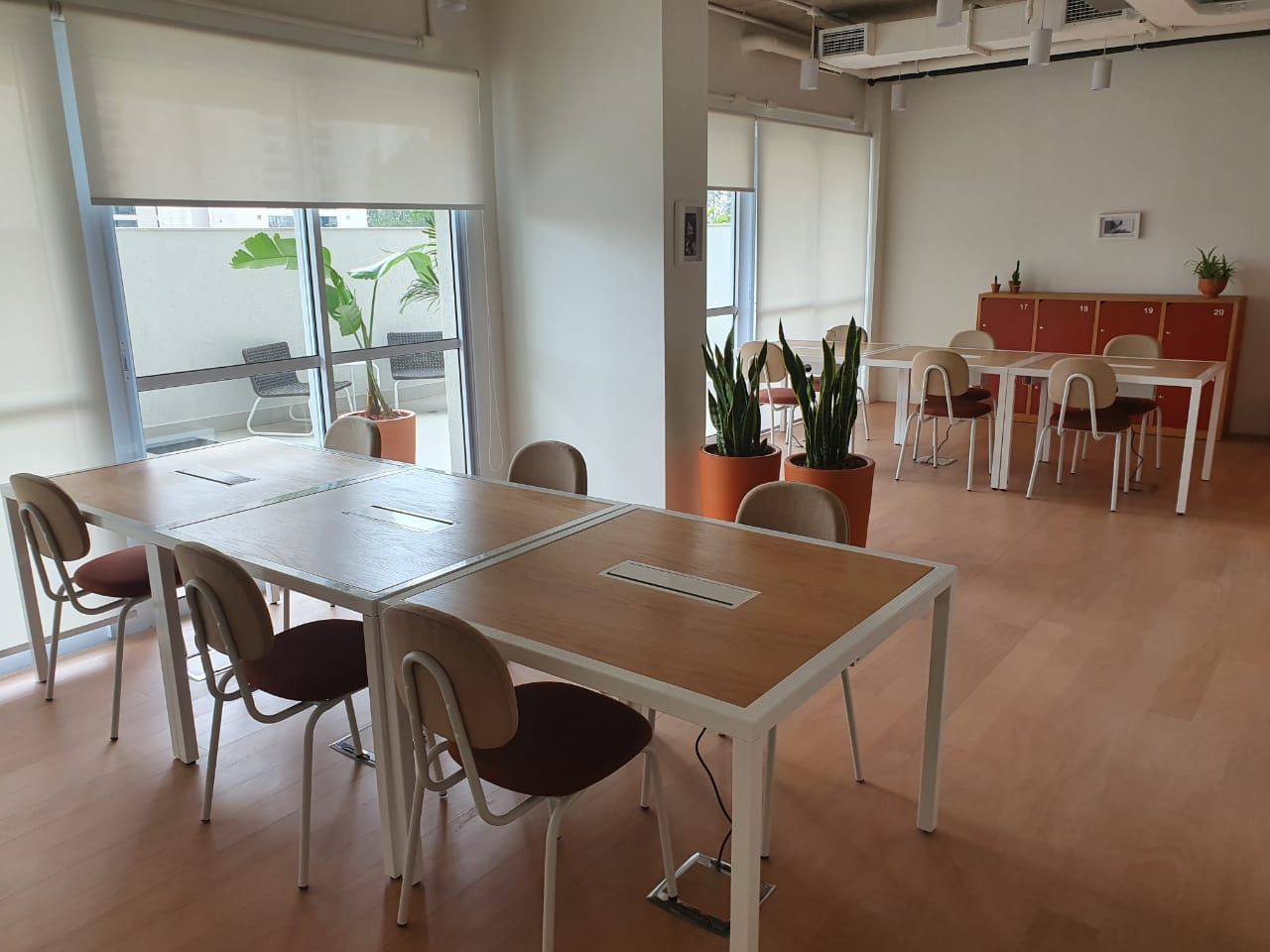 Sala de reuniões iluminada com mesas, cadeiras, vasos de plantas e uma pequena área de refeições junto a grandes janelas.