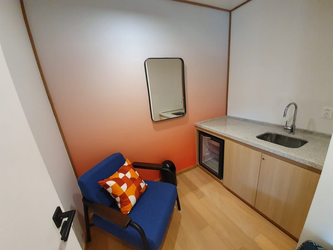 Pequena sala de espera com cadeira azul, almofada laranja, pia, armário e espelho de parede.