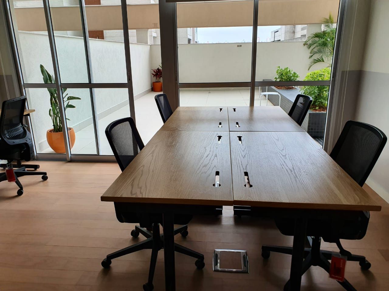 Sala de conferências moderna com uma longa mesa de madeira, cadeiras pretas e grandes janelas com vista para a vegetação.