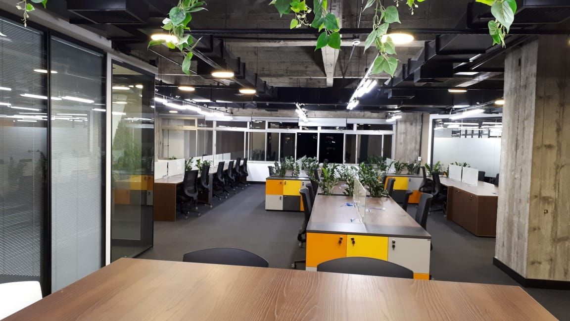 Escritório em plano aberto com mesas, cadeiras, plantas e iluminação aparente no teto.