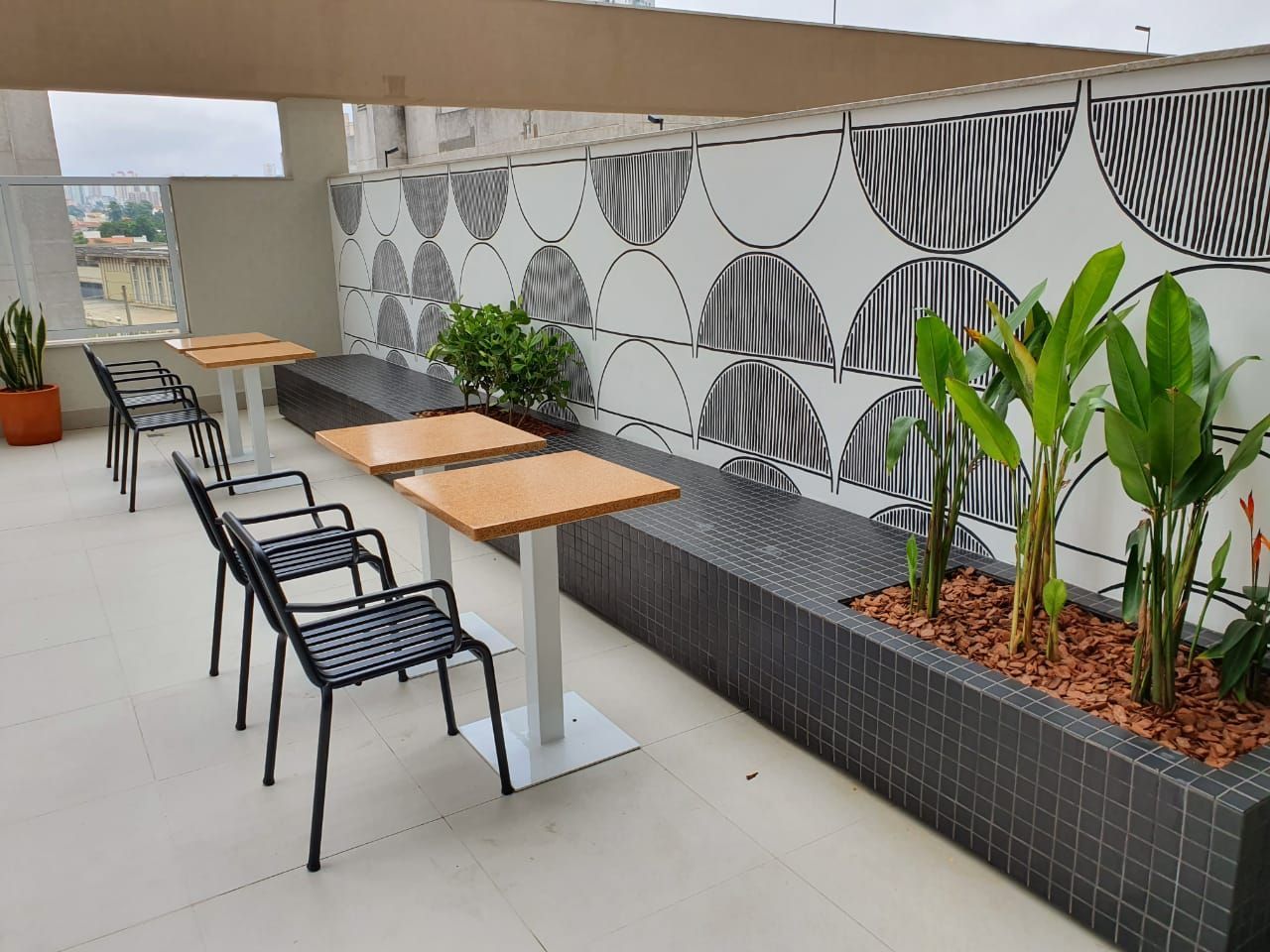 Área externa com mesas e cadeiras pretas, mesas de madeira e vasos de plantas ao lado de uma parede estampada.