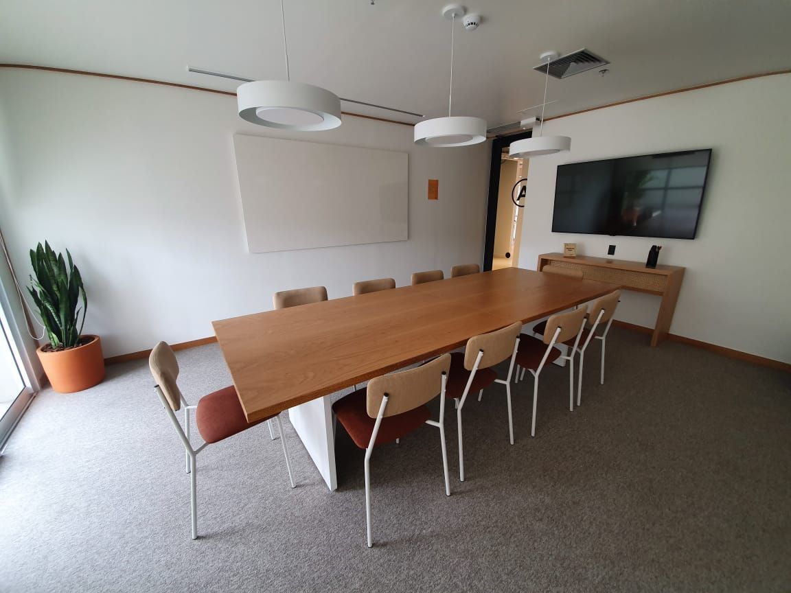 Sala de conferências moderna com mesa comprida de madeira, cadeiras brancas, TV de parede e planta em vaso.