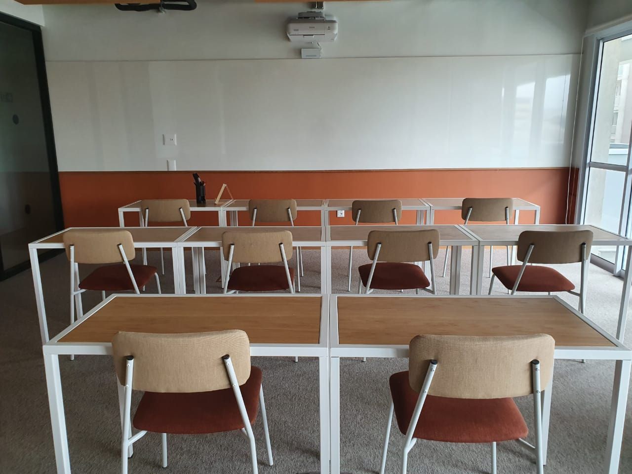 Sala de aula vazia com fileiras de carteiras e cadeiras voltadas para um quadro branco, parede com detalhes em laranja e janelas à direita.