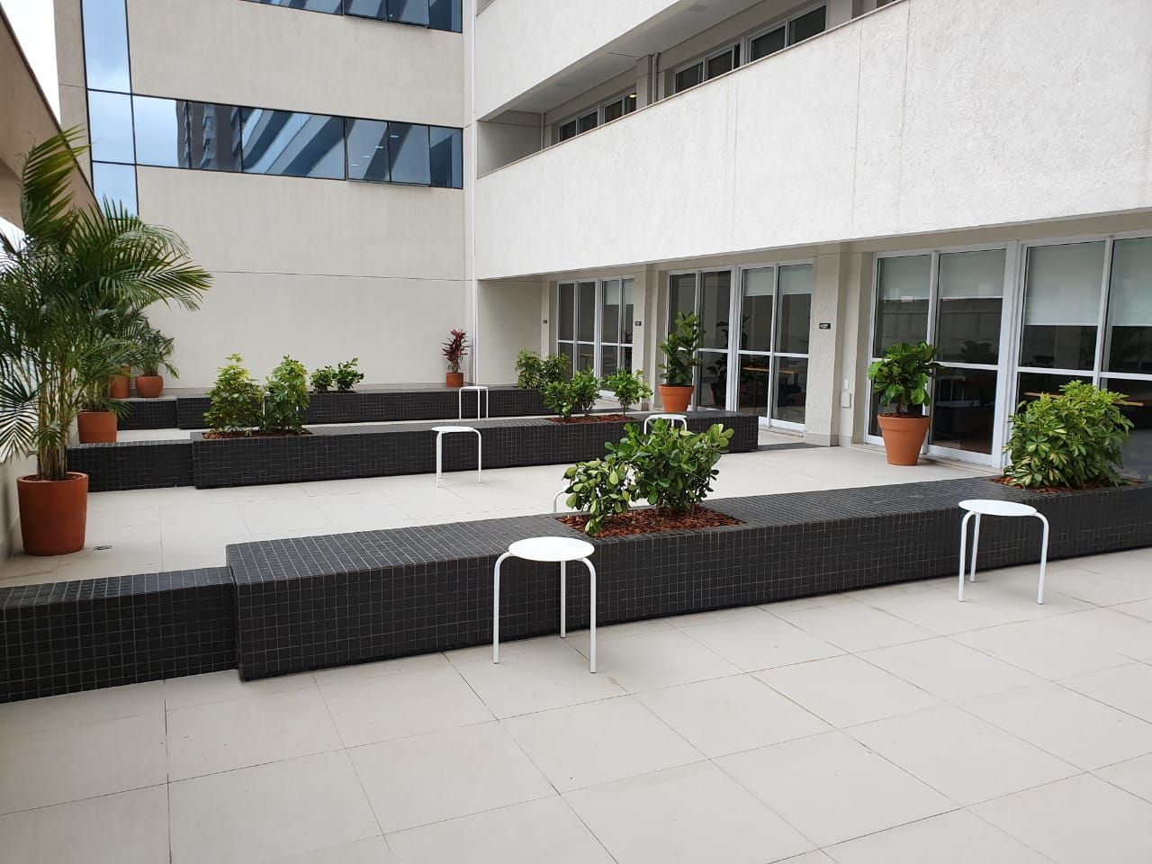 Pátio moderno com bancos pretos em níveis, paredes brancas do edifício, plantas em vasos e piso de azulejos.