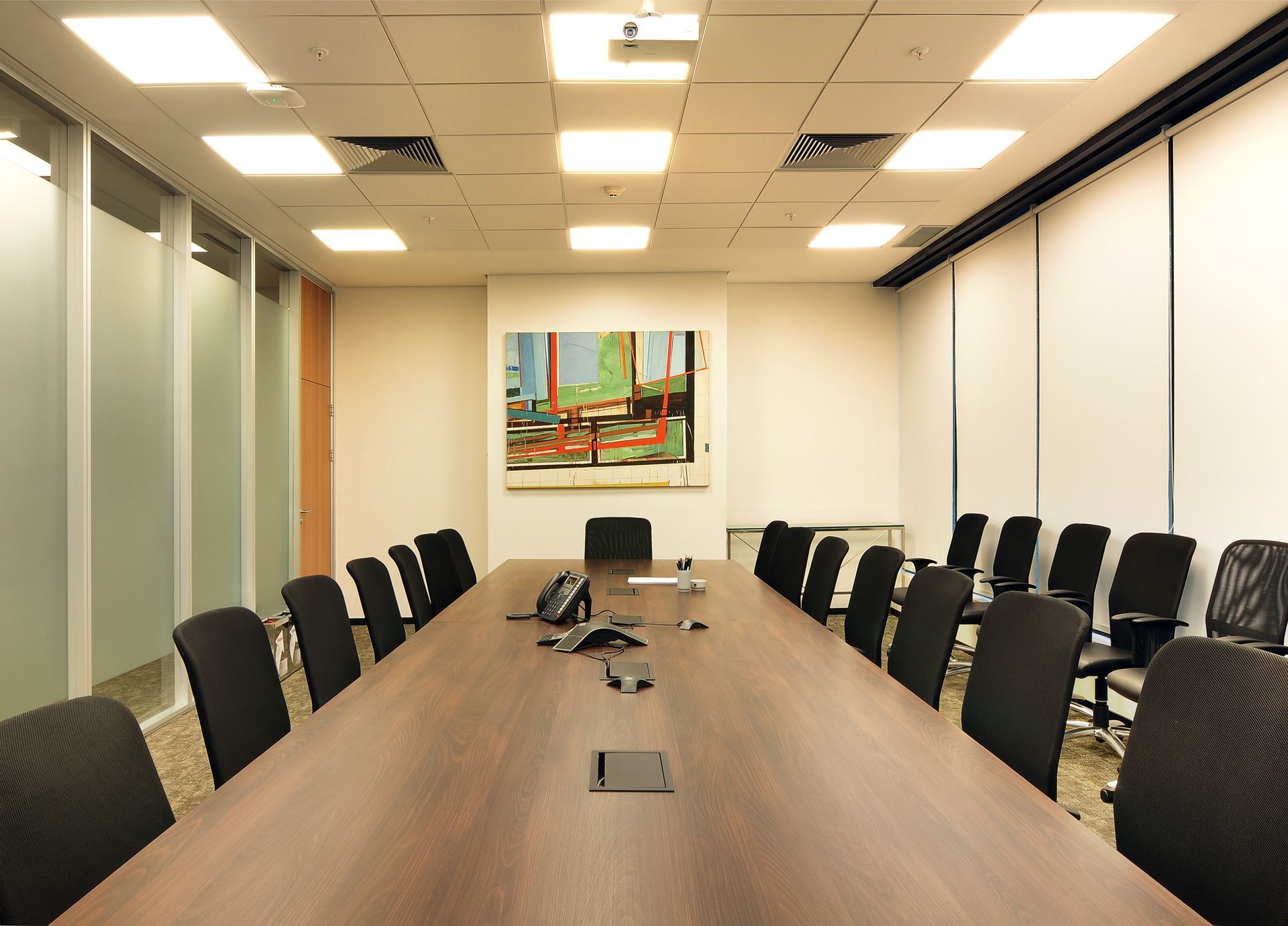 Sala de conferências vazia com uma longa mesa de madeira, cadeiras pretas, luzes no teto e um quadro na parede.