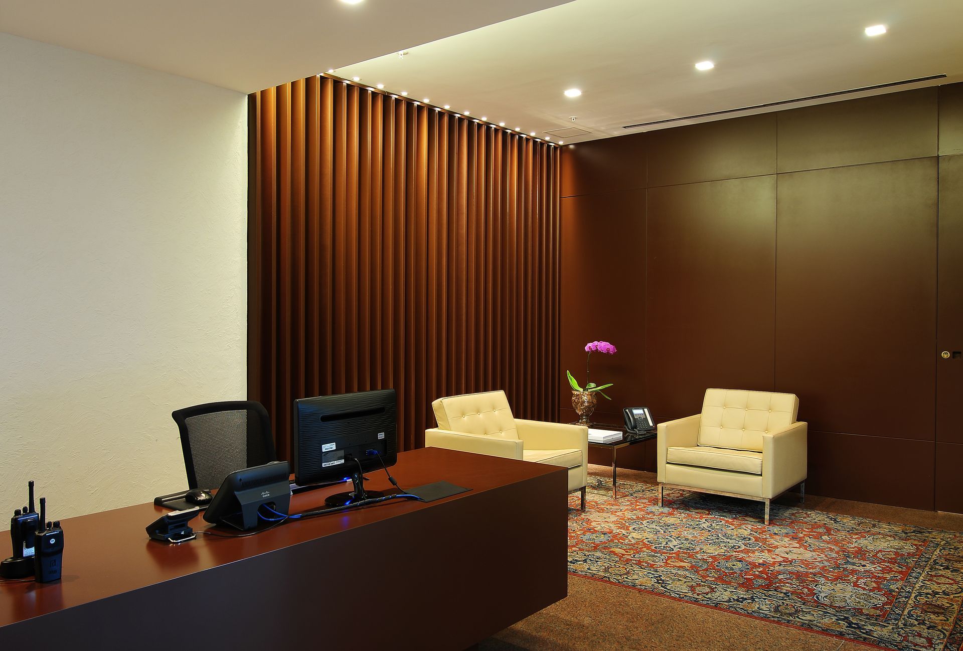 Área de recepção de escritório moderna com mesa, cadeiras e paredes revestidas de madeira em tons quentes.
