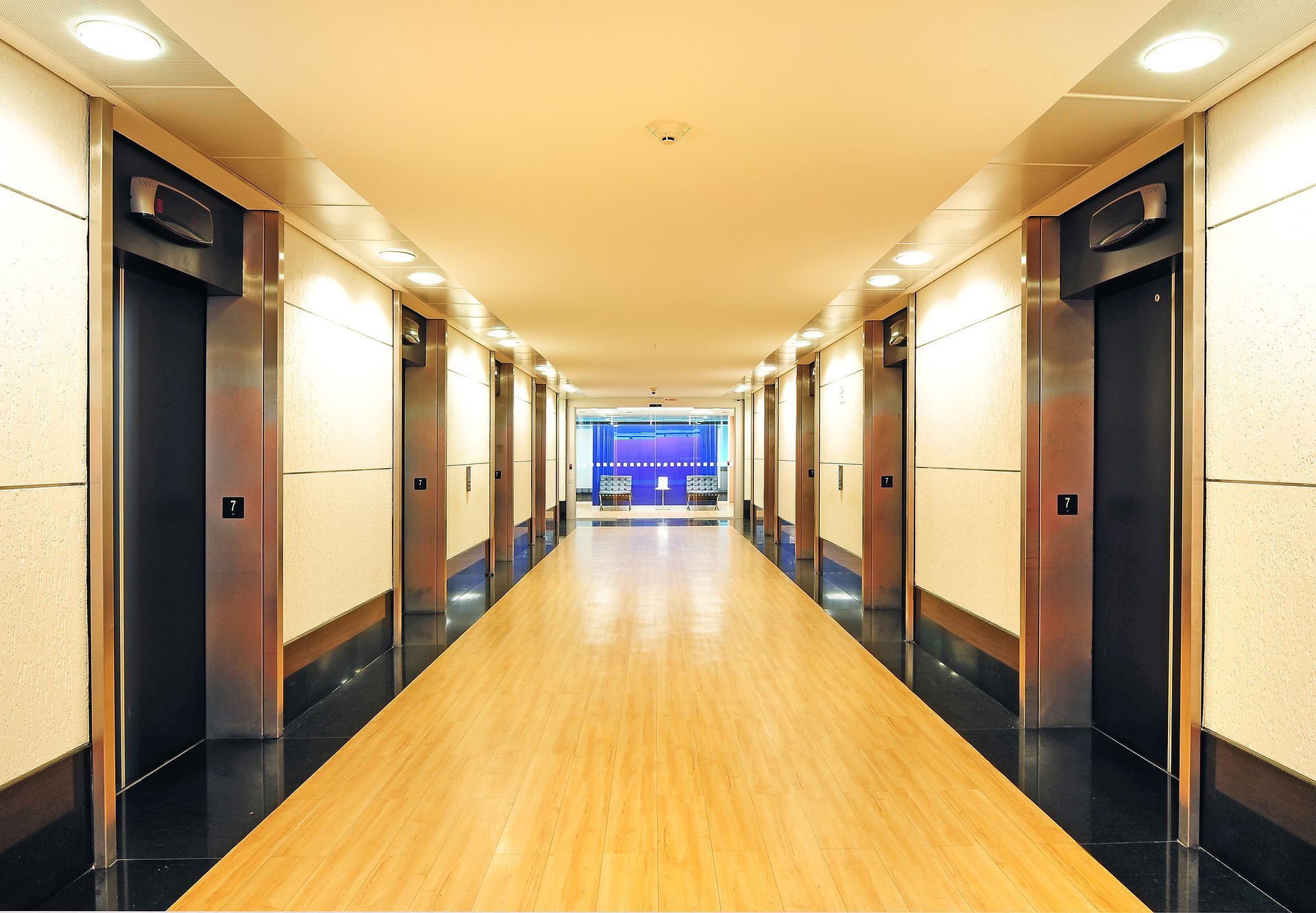 Corredor de hotel iluminado com piso de madeira, portas de elevador escuras e saída com iluminação azul no fundo.