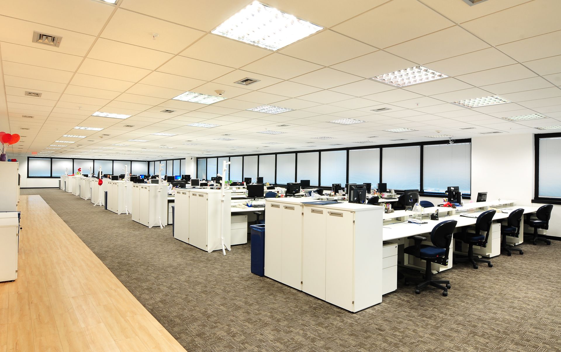 Escritório em plano aberto com fileiras de mesas, computadores e cubículos sob luzes de teto brilhantes.