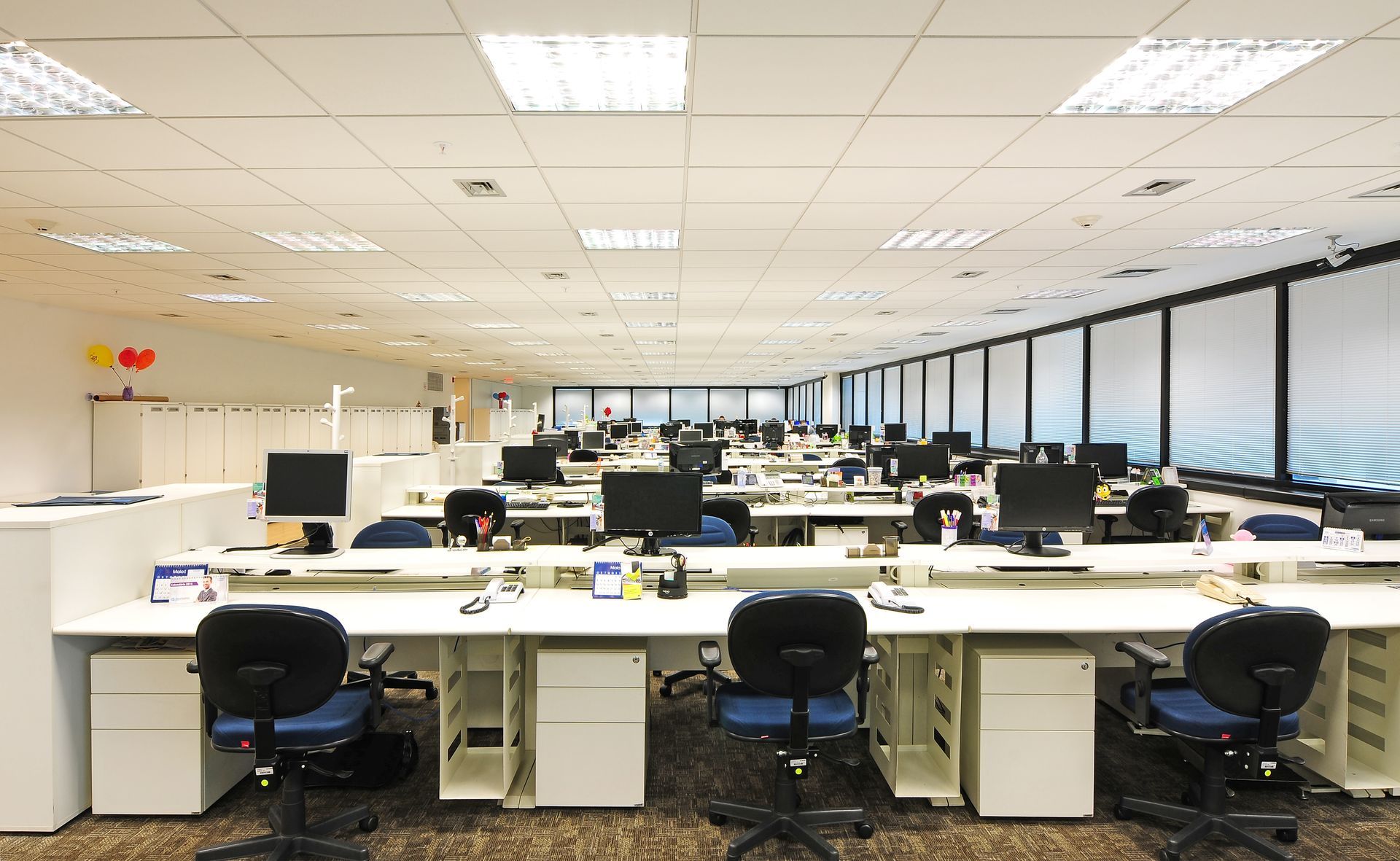 Escritório de planta aberta com fileiras de mesas, computadores e iluminação fluorescente no teto.