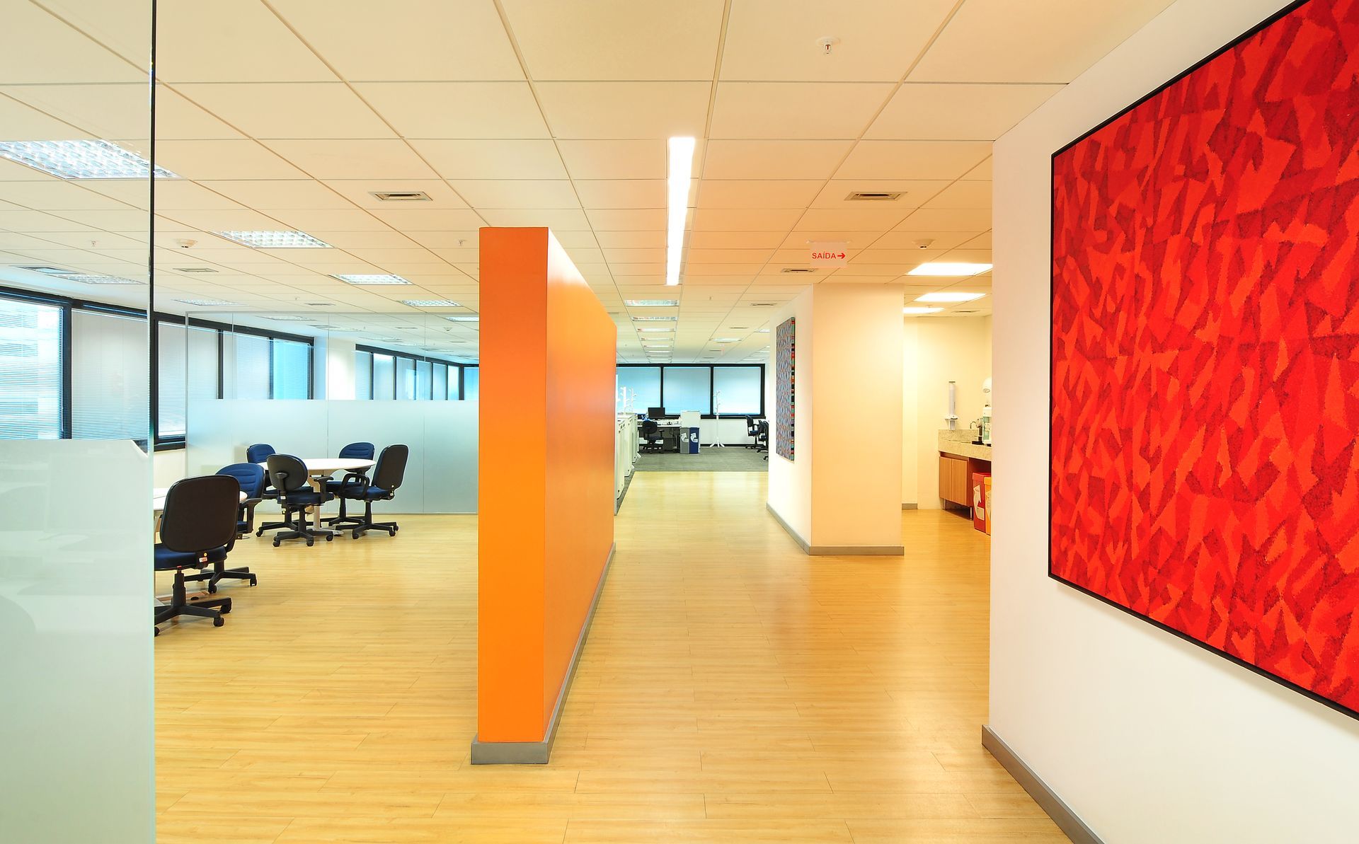 Corredor de escritório iluminado com coluna laranja, arte de parede vermelha e área de trabalho aberta com assentos ao fundo.