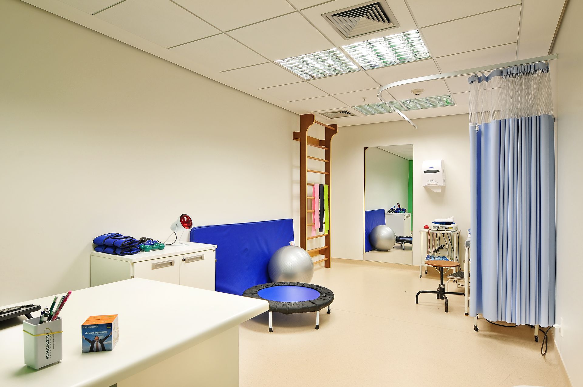 Sala de tratamento hospitalar com cama, cortina para privacidade, equipamentos médicos e mesa de exame.