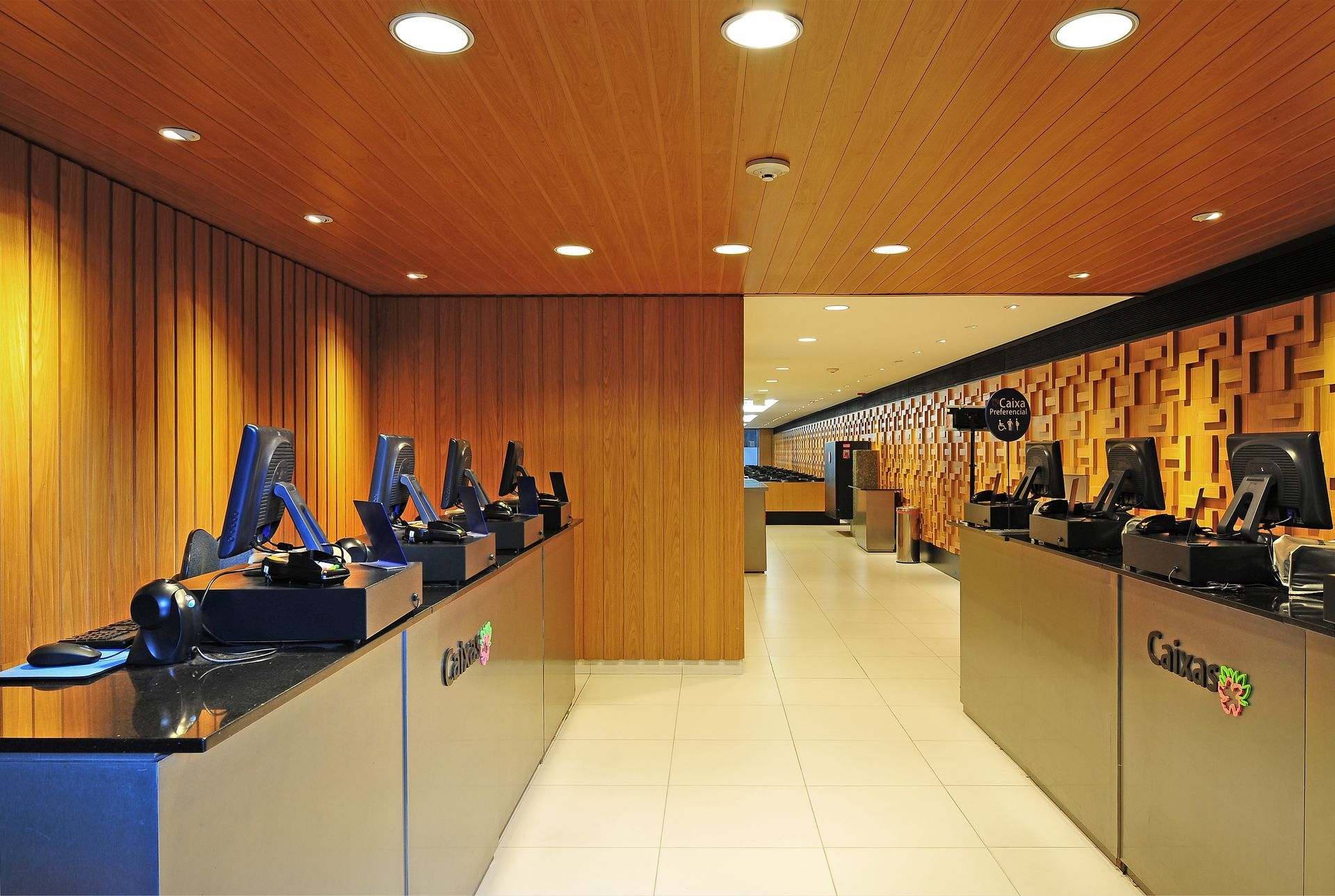 Área de recepção moderna com paredes revestidas de madeira, piso branco e estações de computador em ambos os lados.