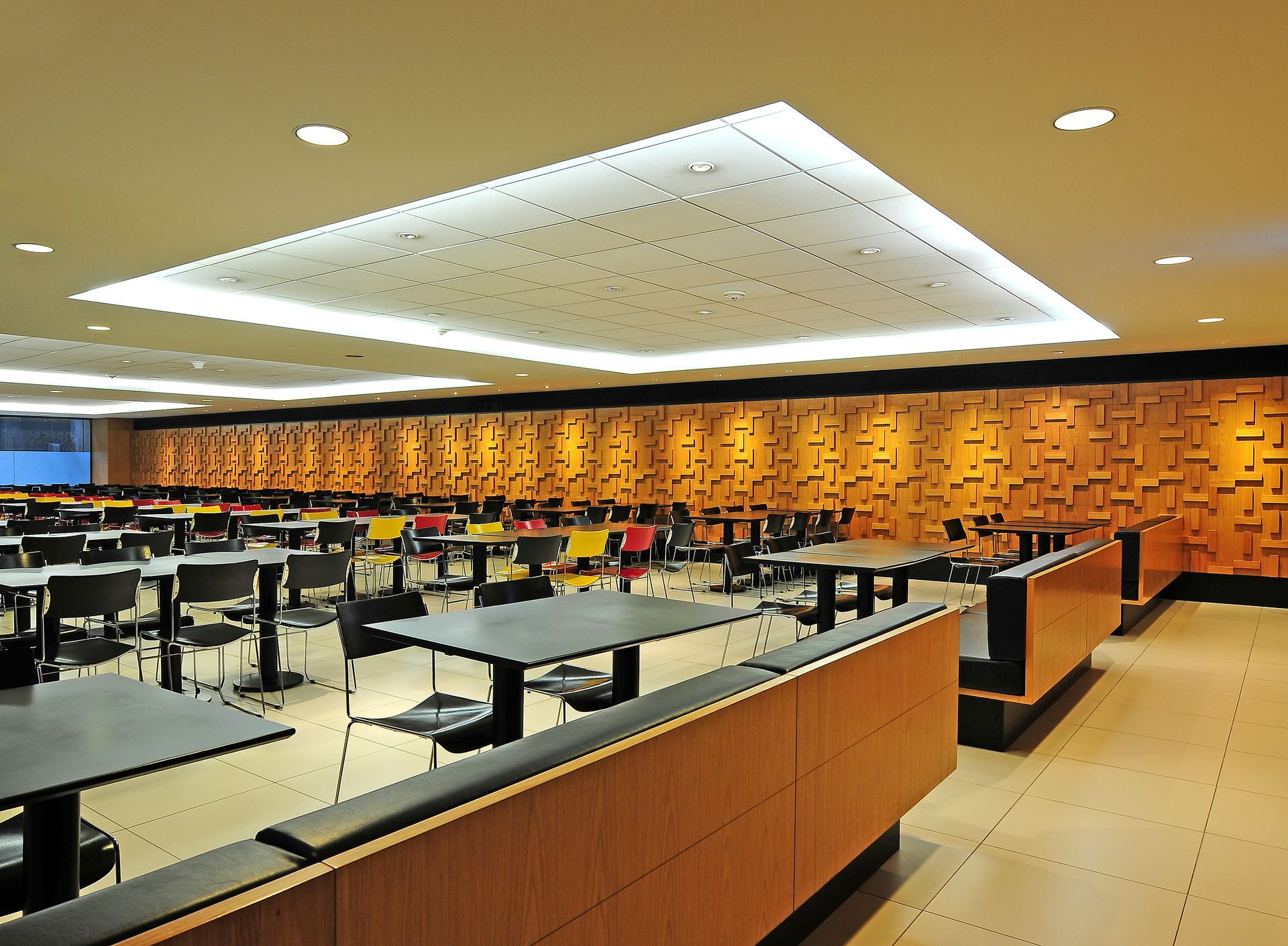 Cafeteria moderna e vazia, com fileiras de mesas, cadeiras bege e painéis de parede em um tom quente de laranja, sob luzes brilhantes no teto.