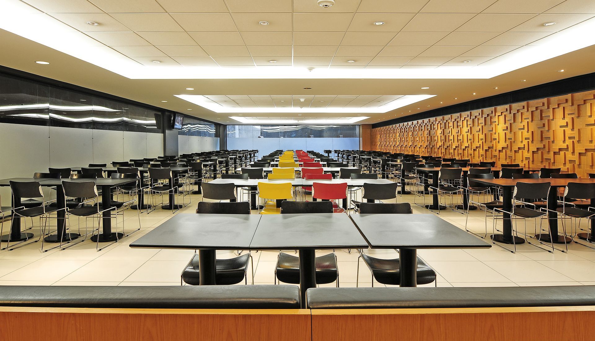 Cafeteria moderna vazia com fileiras de mesas e cadeiras, mesas centrais com bandejas vermelhas e amarelas.