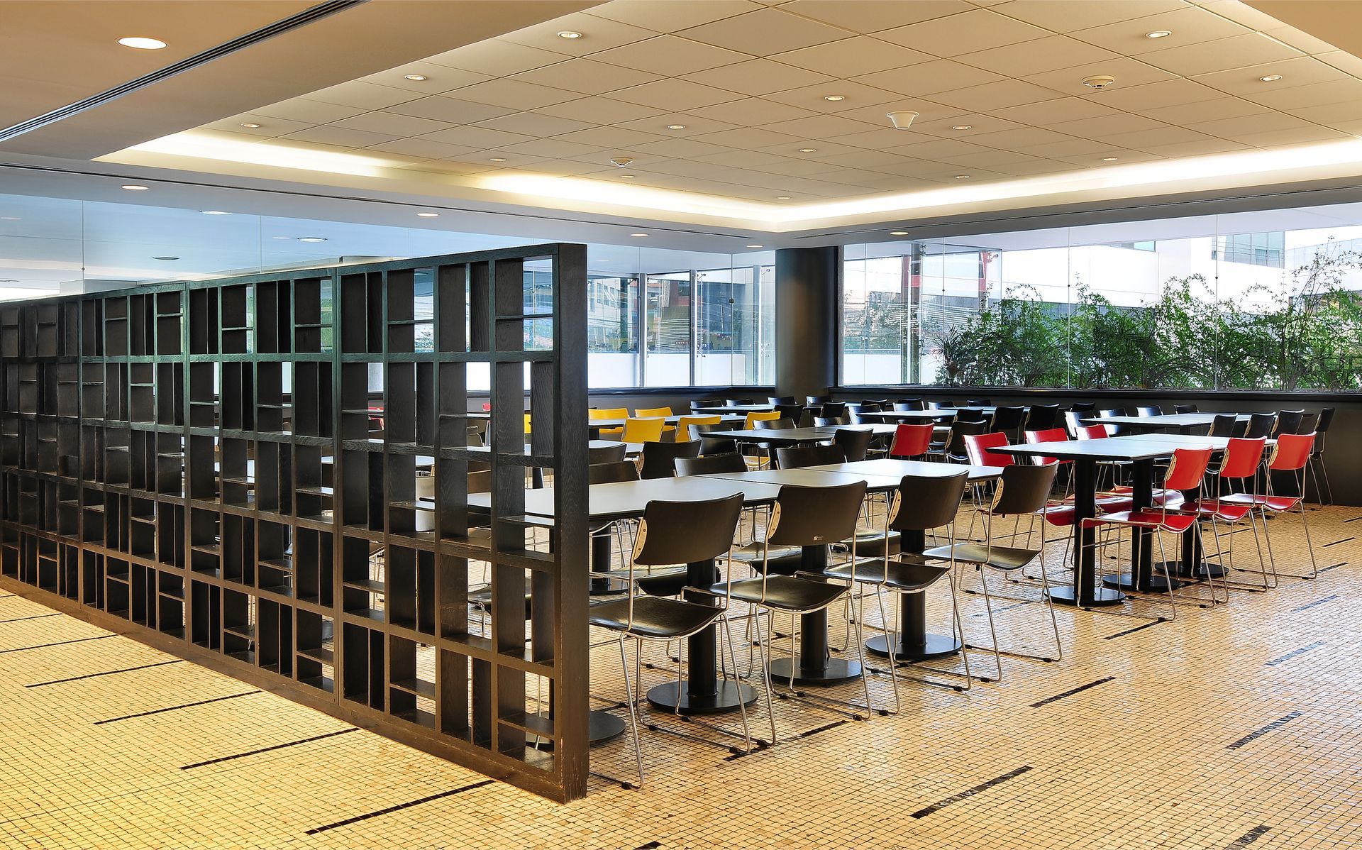 Área de estudo moderna de biblioteca com estantes de livros, mesas e cadeiras vermelhas e pretas sob luzes brilhantes no teto.