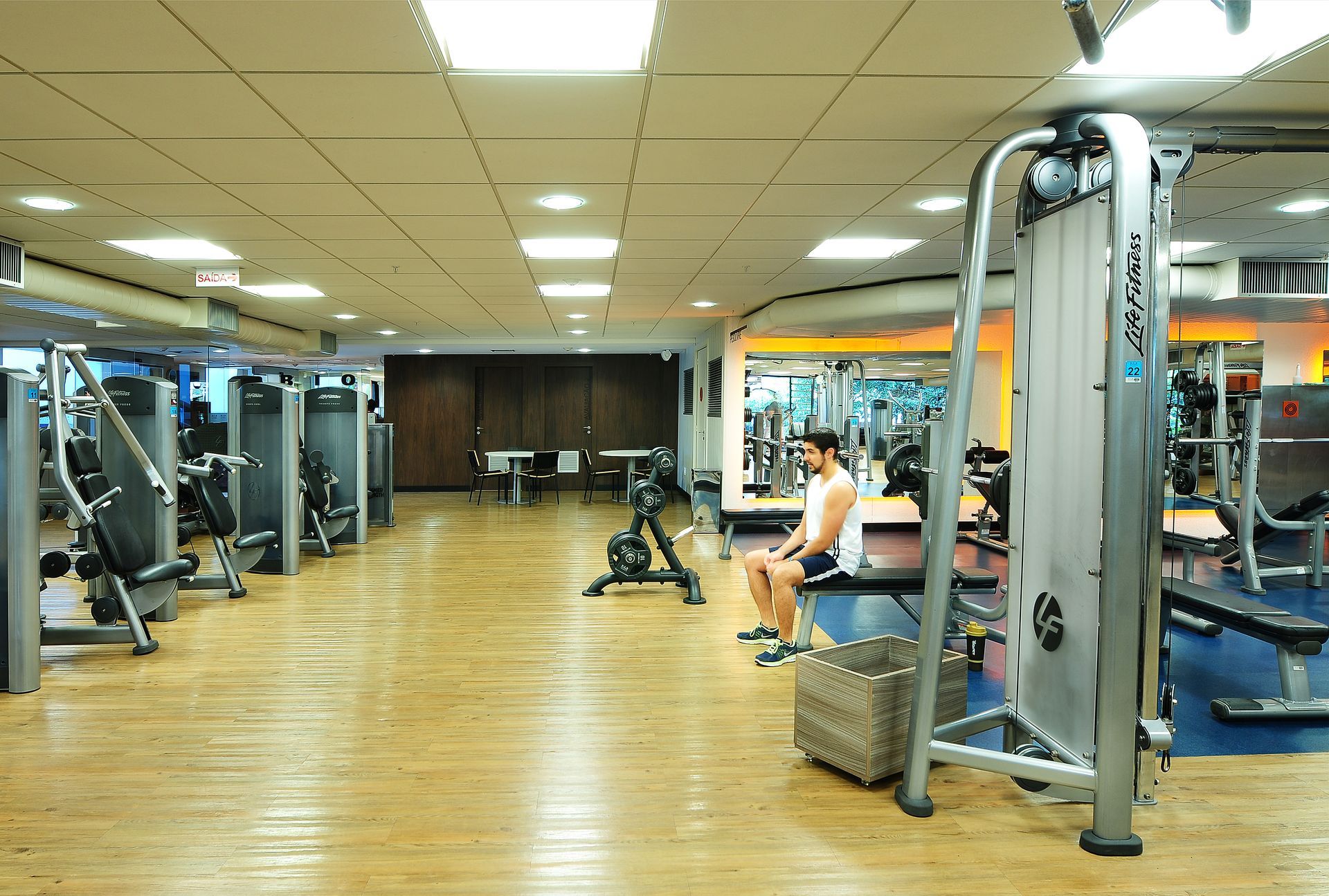 Interior luminoso de um ginásio com piso de madeira, aparelhos de musculação e uma pessoa a exercitar-se ao fundo.