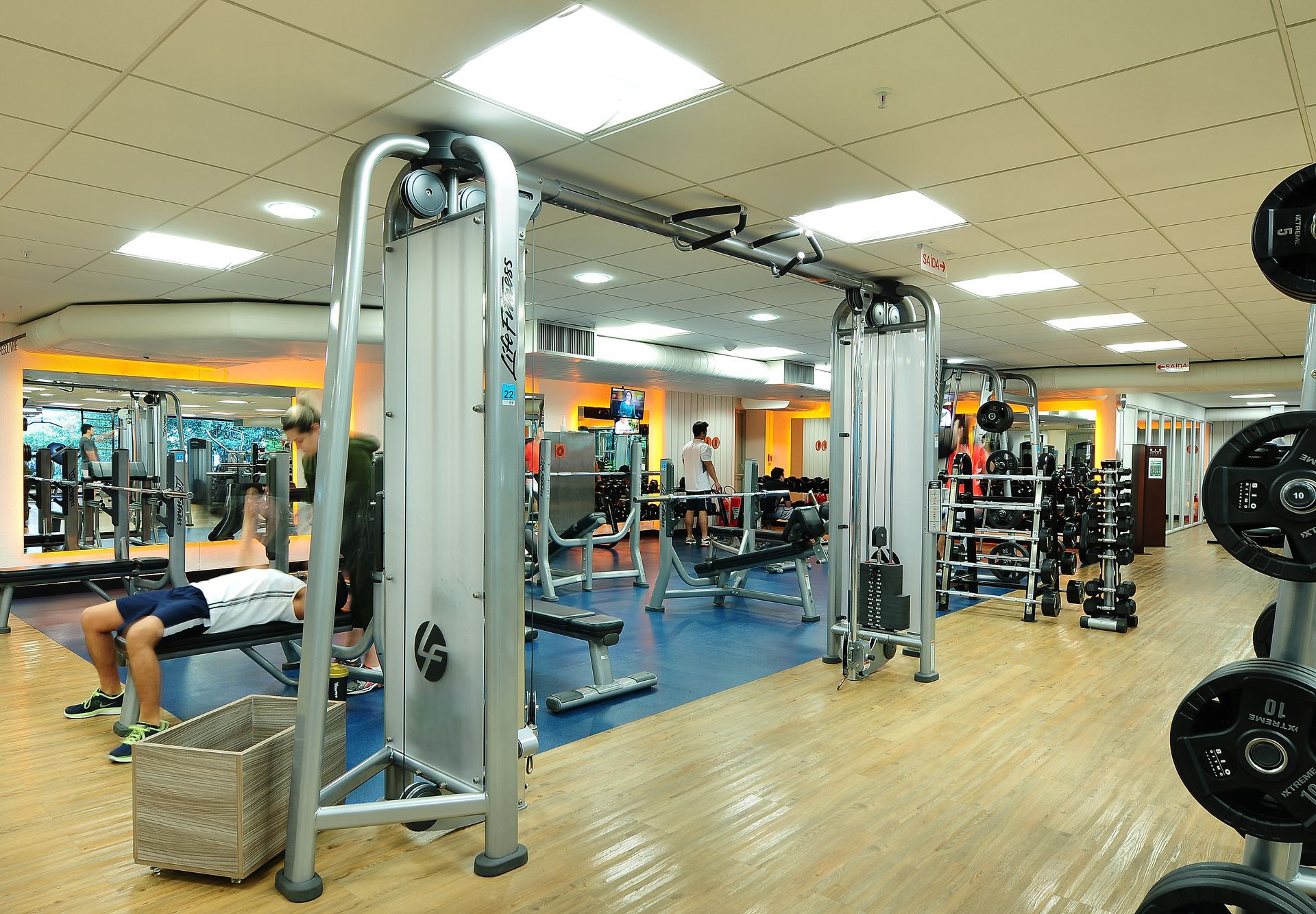 Interior de academia com aparelhos de musculação, espelhos e uma pessoa se exercitando em um colchonete.