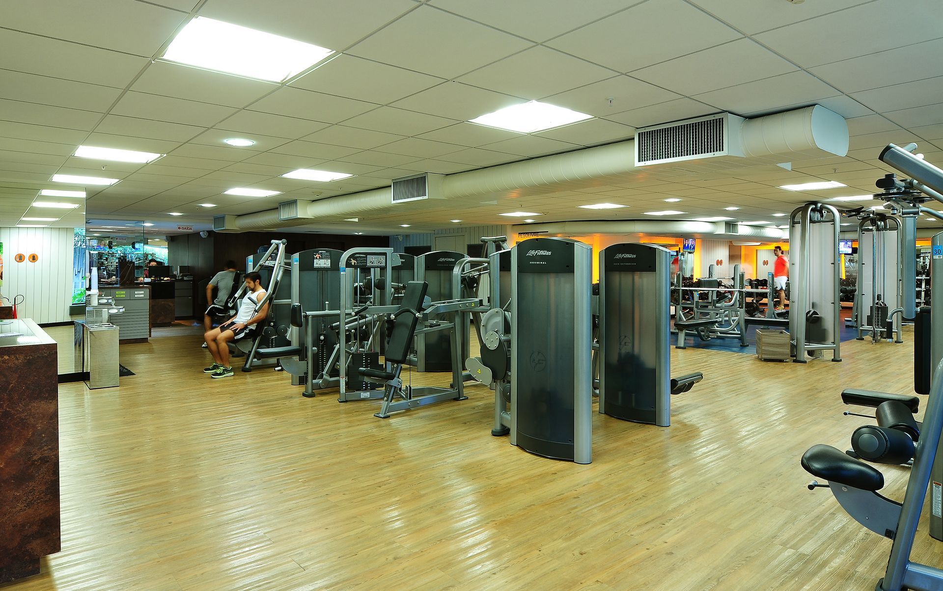 Academia interna com aparelhos de ginástica, espelhos e algumas pessoas se exercitando em um piso de madeira.