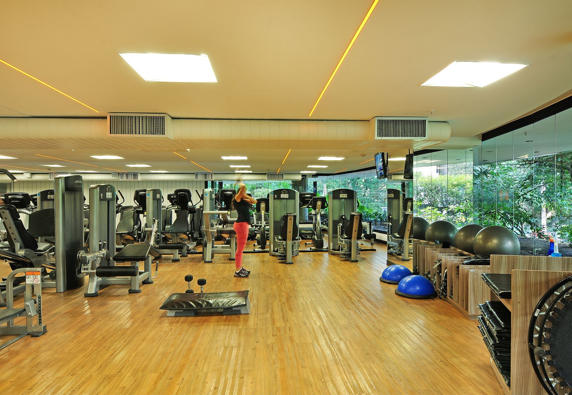 Academia iluminada com paredes espelhadas, piso de madeira, equipamentos de ginástica e uma pessoa se alongando vestida de vermelho perto do centro.