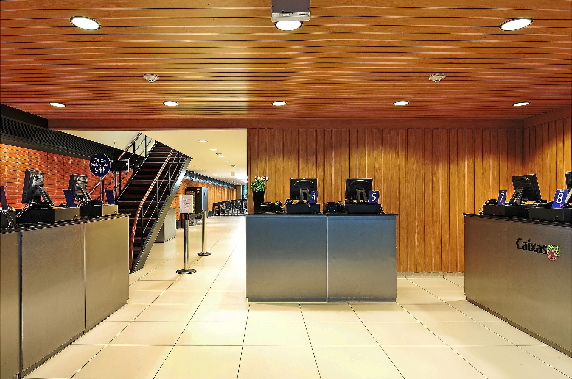 Área de recepção moderna com teto de madeira, piso de azulejo, balcões e computadores.