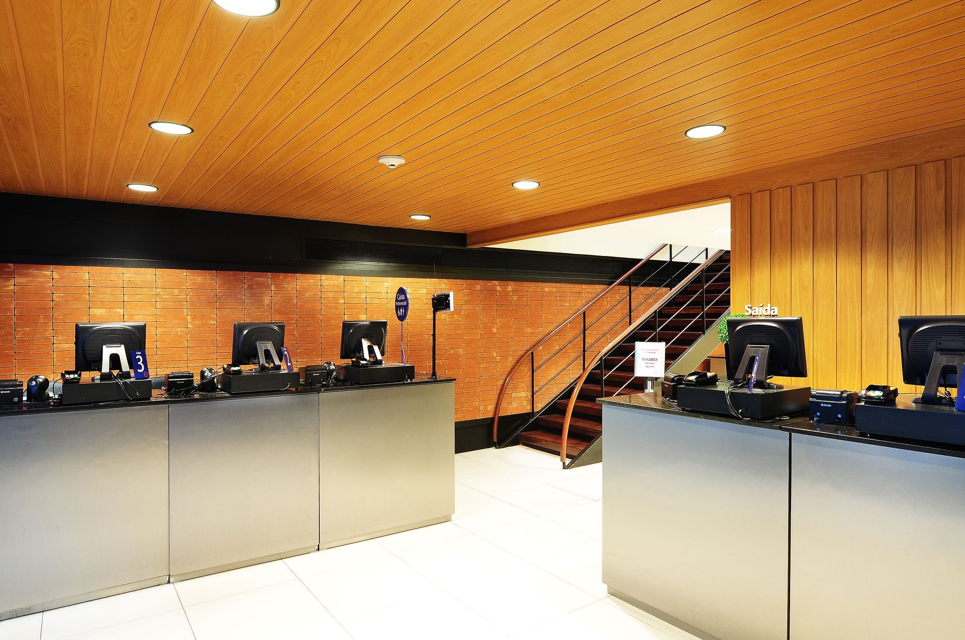 Balcão de atendimento com computadores em uma sala com painéis de madeira, com vários terminais e uma escada central.