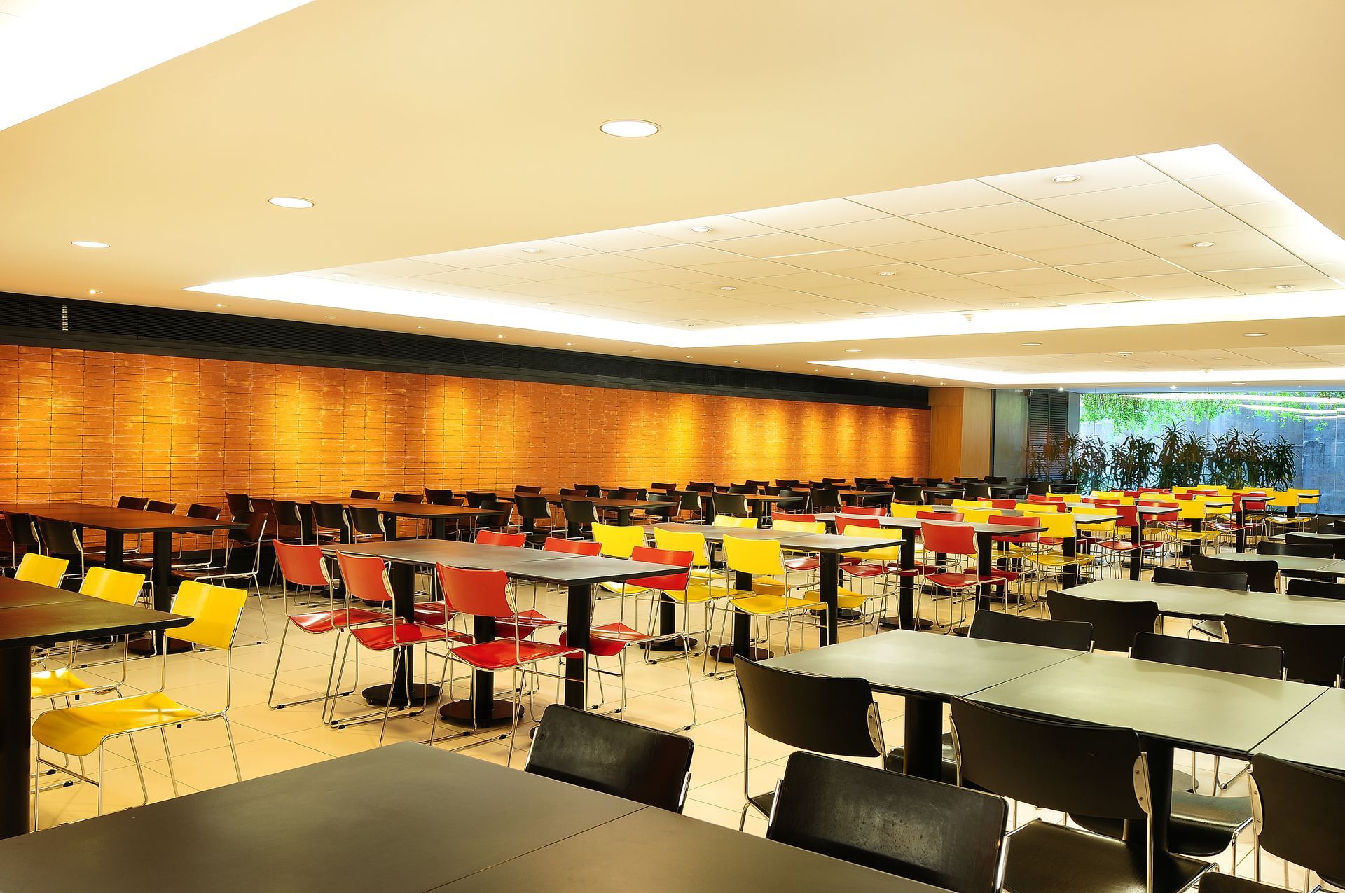 Cafeteria moderna e vazia, com cadeiras e mesas pretas, vermelhas e amarelas, sob luzes de teto aconchegantes.