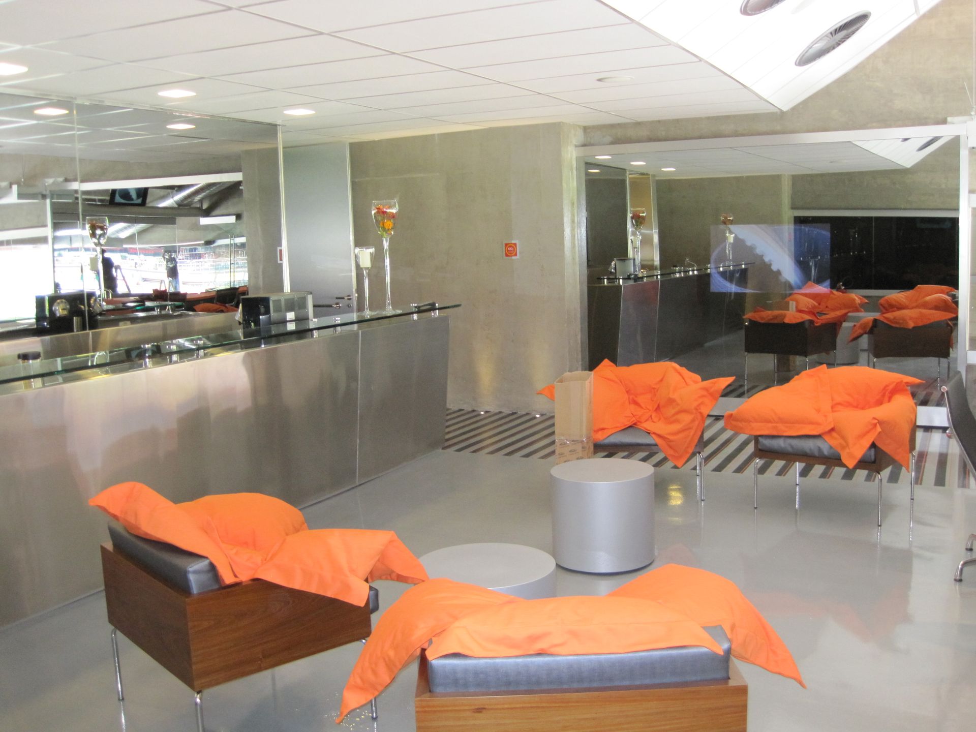 Sala de estar moderna com cadeiras laranja, mesas cinza e piso polido em um saguão de escritório iluminado.