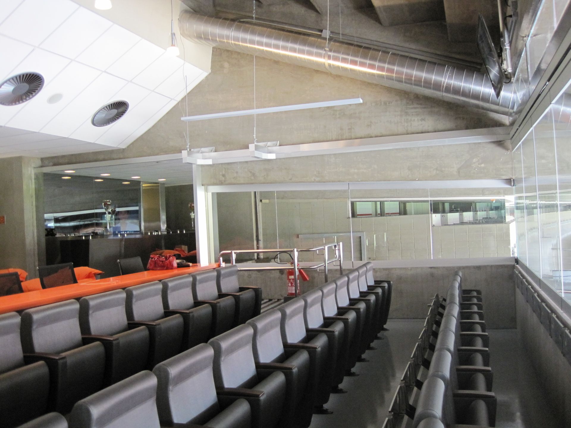 Auditório moderno vazio com fileiras de assentos cinza, detalhes em laranja e dutos de teto expostos.