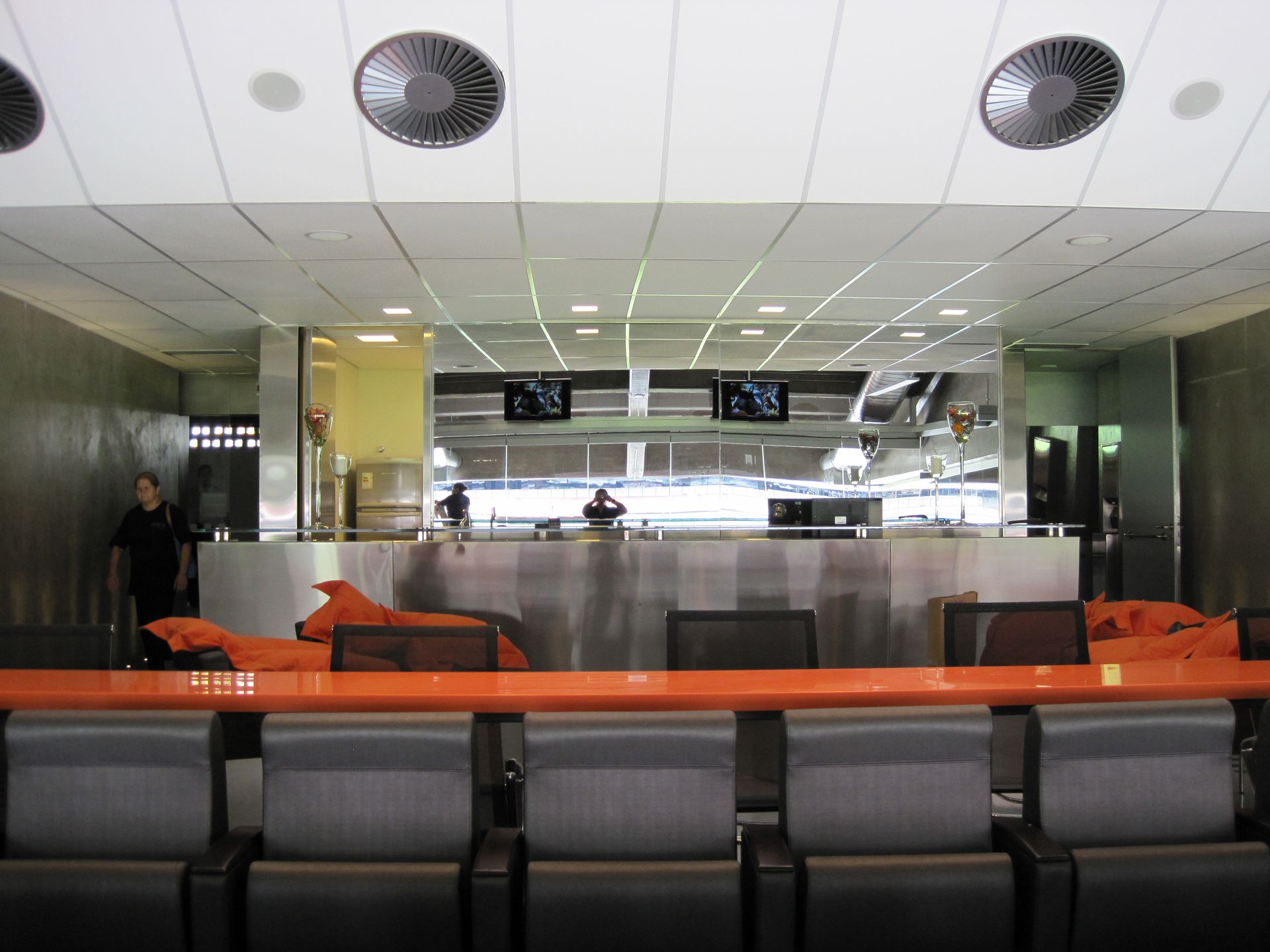 Sala de espera moderna do aeroporto com sofás laranja, cadeiras cinza e um bar com vista panorâmica de vidro.
