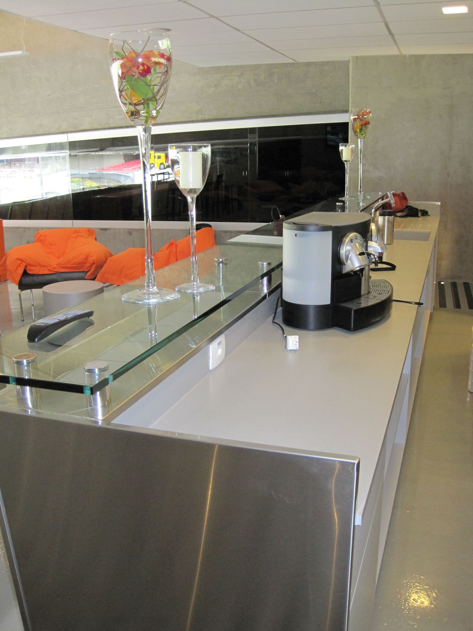 Sala de estar moderna com cadeiras laranja, guarda-corpo de vidro e estação de café em uma bancada.