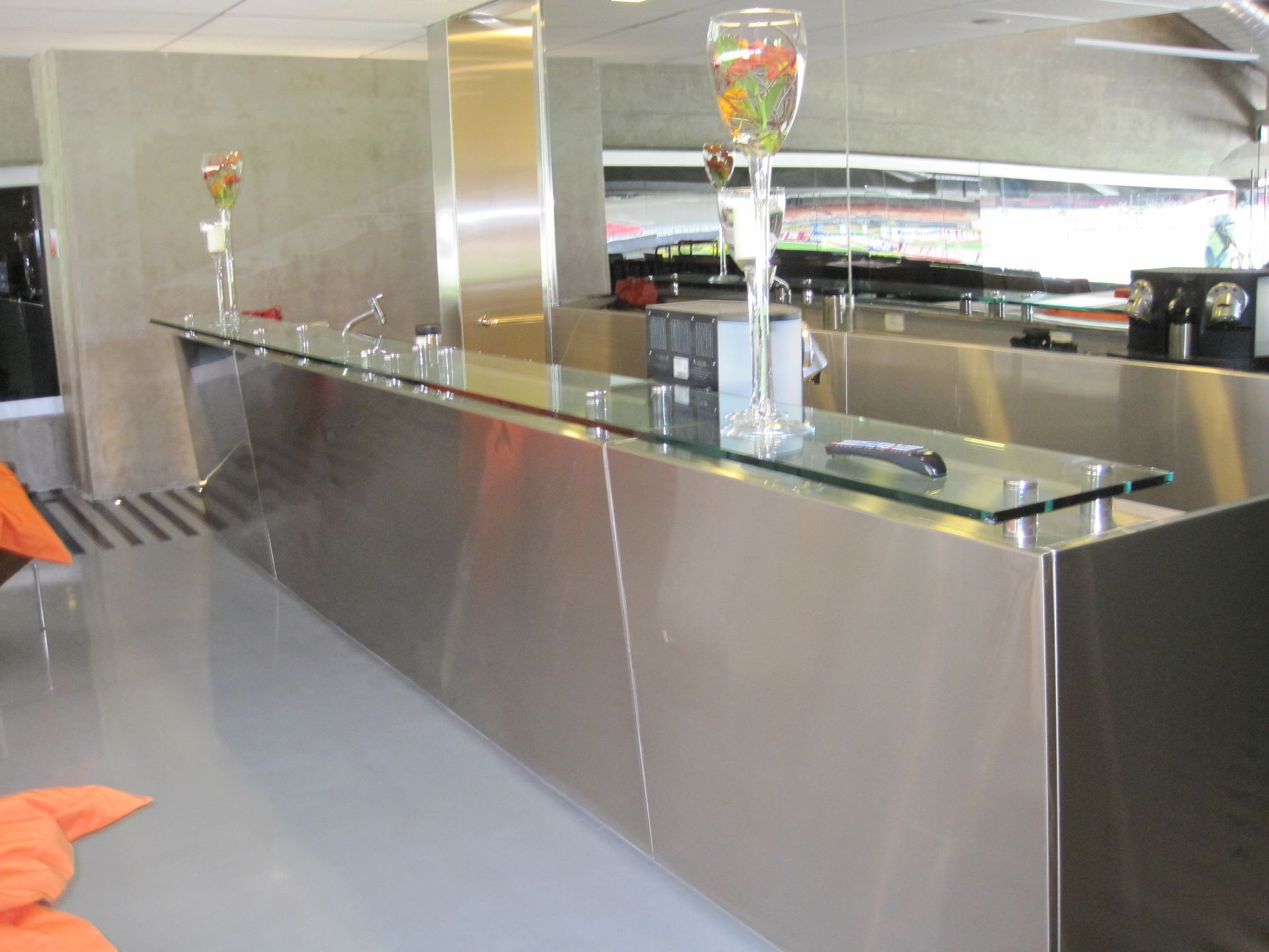 Cozinha moderna com uma longa bancada de aço inoxidável e tampo de vidro, além de assentos laranja e plantas suspensas.