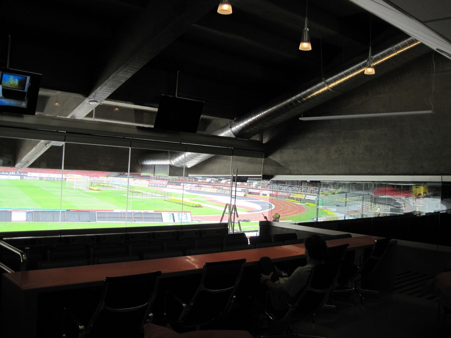 Camarote coberto com vista para um campo de futebol iluminado e com assentos pretos vazios em primeiro plano.