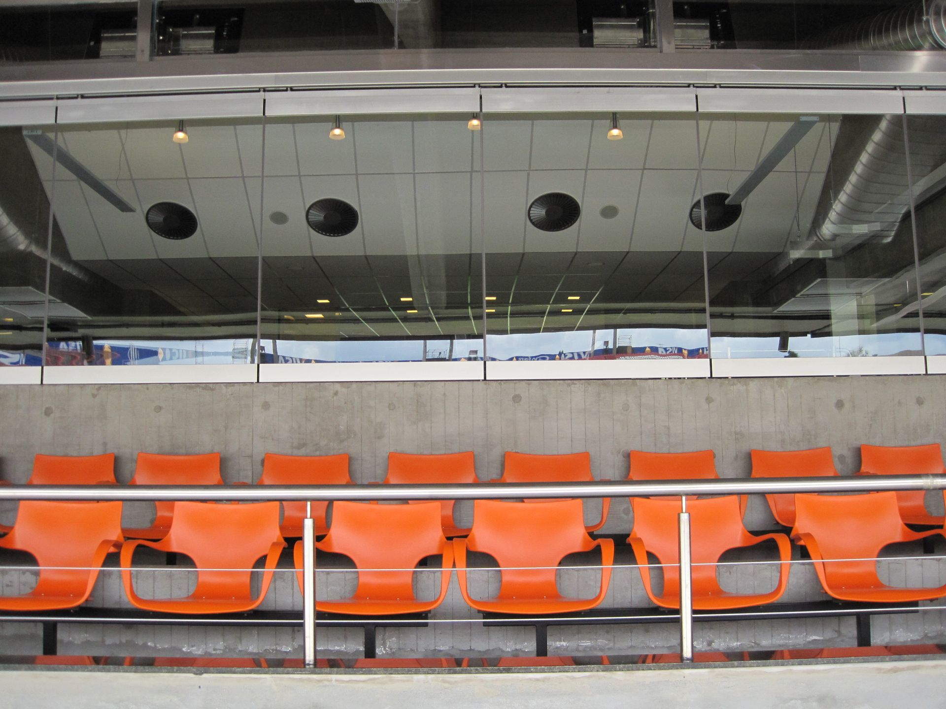 Assentos vazios de estádio na cor laranja, sob uma arquibancada coberta com fachada de vidro.