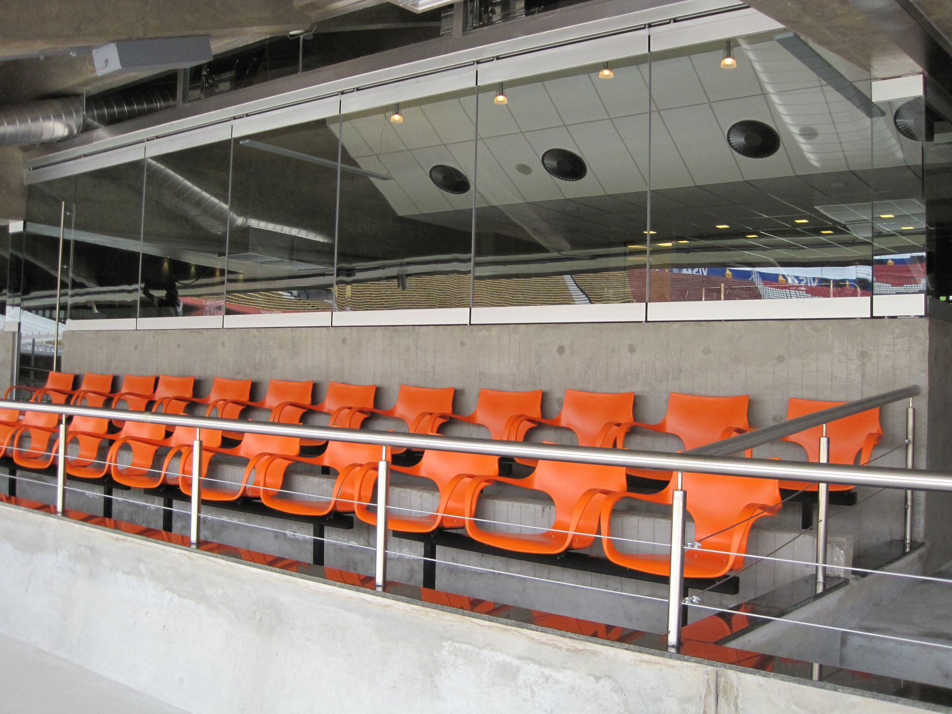 Fileira de assentos vazios na cor laranja, atrás de uma barreira de vidro, em um moderno estádio esportivo.