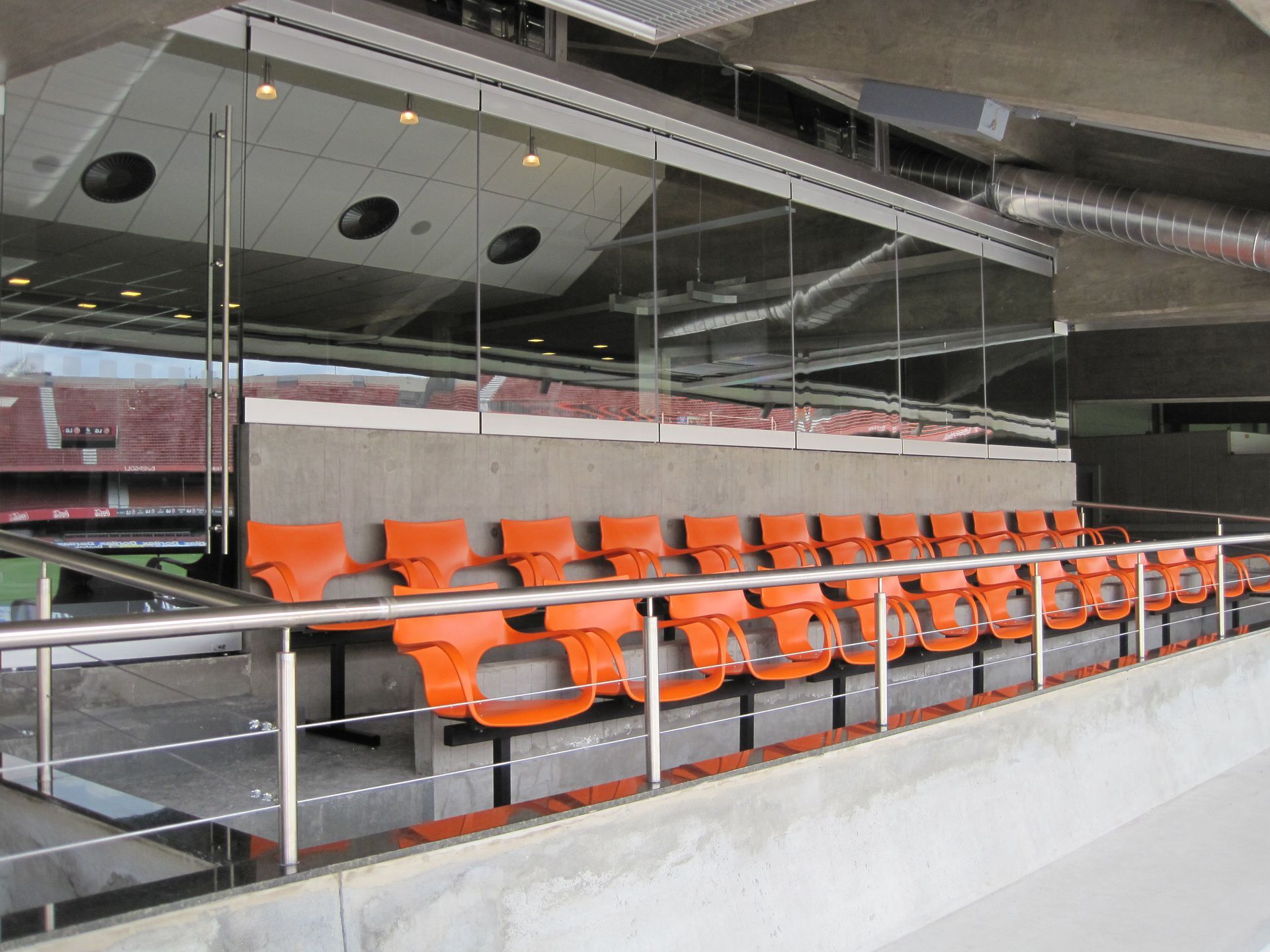 Assentos vazios no estádio com fileiras de cadeiras laranjas atrás de um guarda-corpo de vidro.