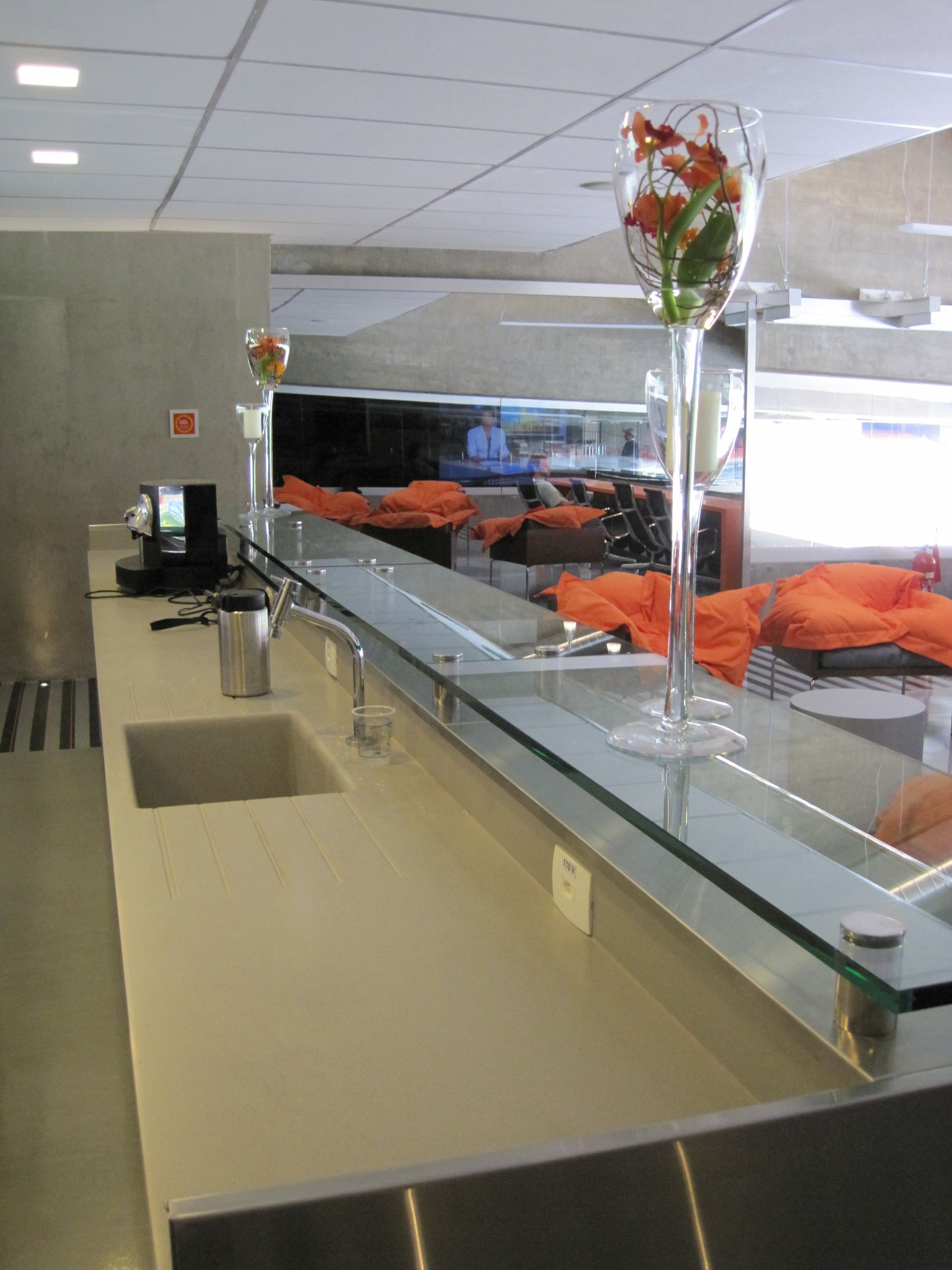 Balcão de cafeteria com bandejas de servir, flores de laranjeira e um protetor de vidro contra espirros em um refeitório iluminado.